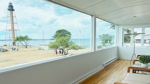 A room with a large window overlooking a beach, a wooden floor, and a table with chairs. - The Sunset Lighthouse Marblehead - Exclusive Waterfront 3brm/2.5bath Home (Marblehead)