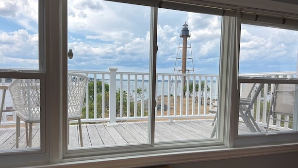 A room with a large window overlooking a wooden deck with white railings and a lighthouse in the distance. - The Sunset Lighthouse Marblehead - Exclusive Waterfront 3brm/2.5bath Home (Marblehead)