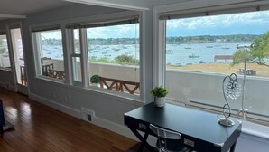 A room with large windows offering a view of a harbor with boats and a beach. - The Sunset Lighthouse Marblehead - Exclusive Waterfront 3brm/2.5bath Home (Marblehead)