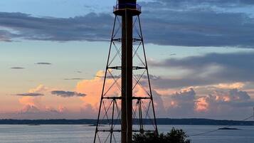 A lighthouse with a metal framework and a light at the top, overlooking a body of water with a sunset in the background.