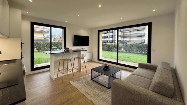 A modern apartment with a kitchen, dining area, and living room. It features large windows, a flat-screen TV, and a sofa. - Jardin privé + parking · 6 pers · plage 2 min (Deauville)