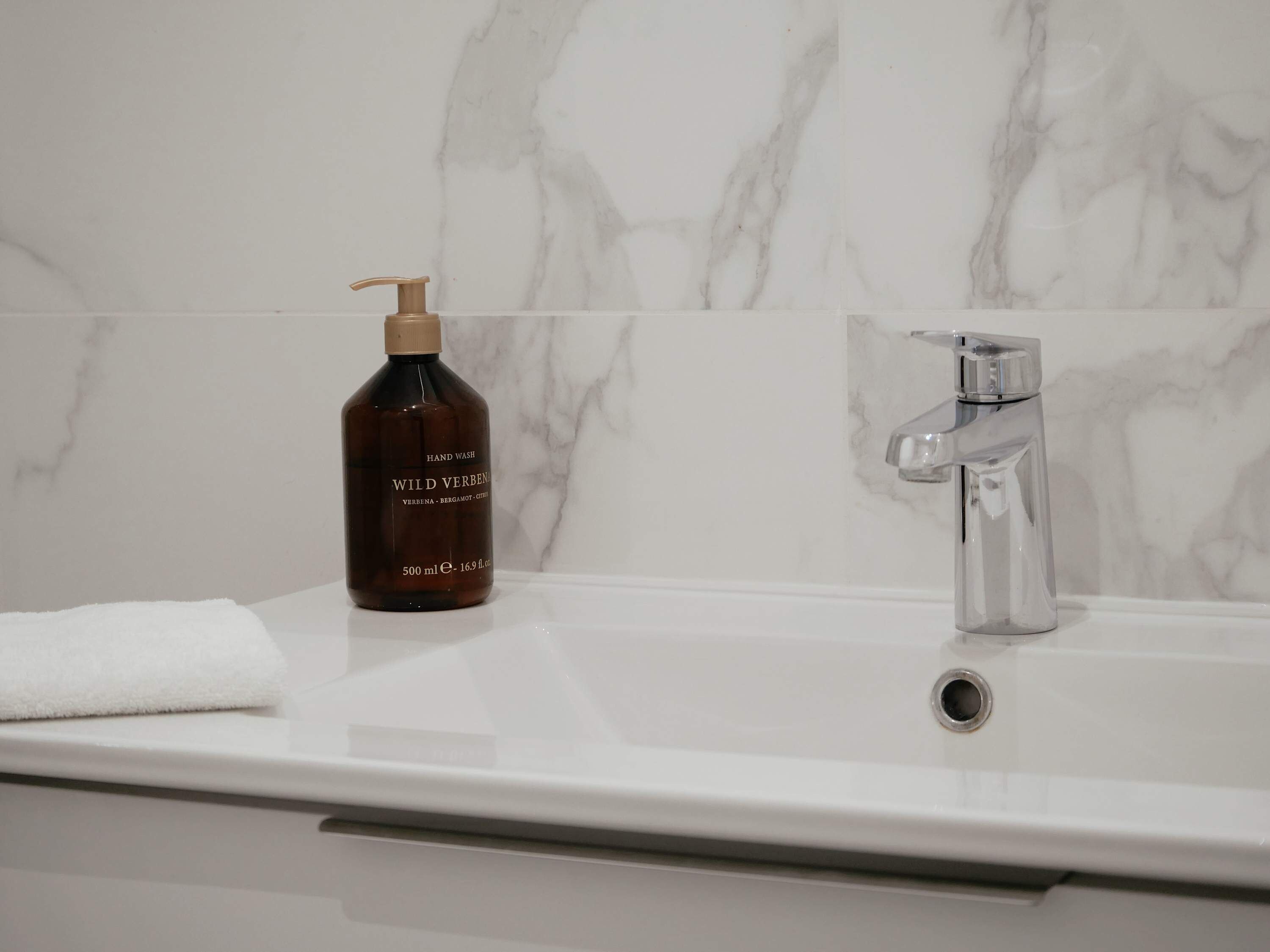 A bathroom with a marble wall, a chrome faucet, and a bottle of Wild Verbena hand wash.