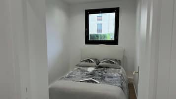 Una habitación estrecha con paredes blancas, una cama al fondo, una ventana con marco negro y vistas a un edificio y follaje exterior.