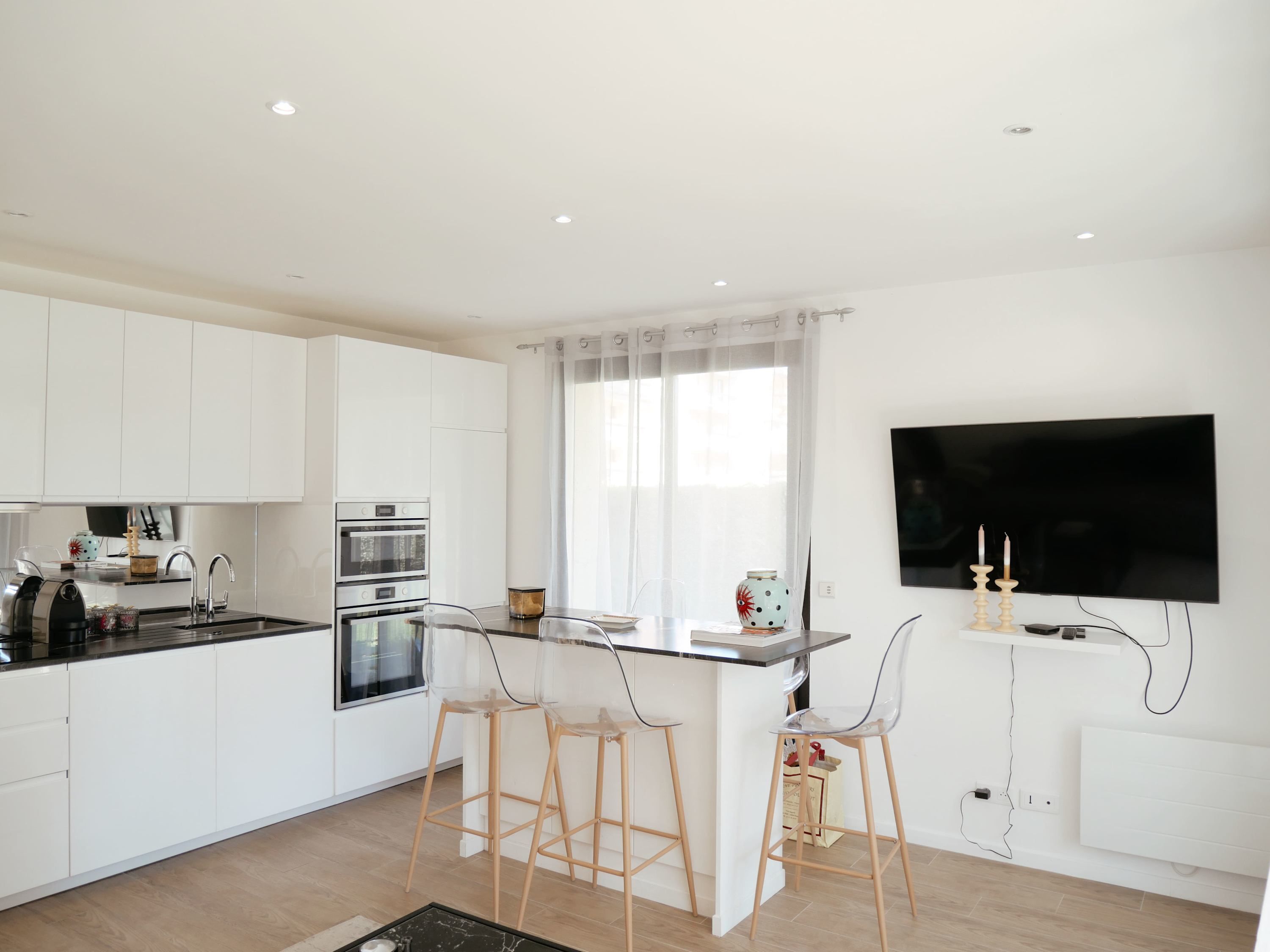 Une cuisine moderne avec des meubles blancs, un îlot central doté de tabourets, un four encastré et un téléviseur à écran plat fixé au mur.