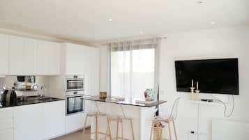 Une cuisine moderne avec des meubles blancs, un îlot central doté de tabourets, un four encastré et un téléviseur à écran plat fixé au mur.