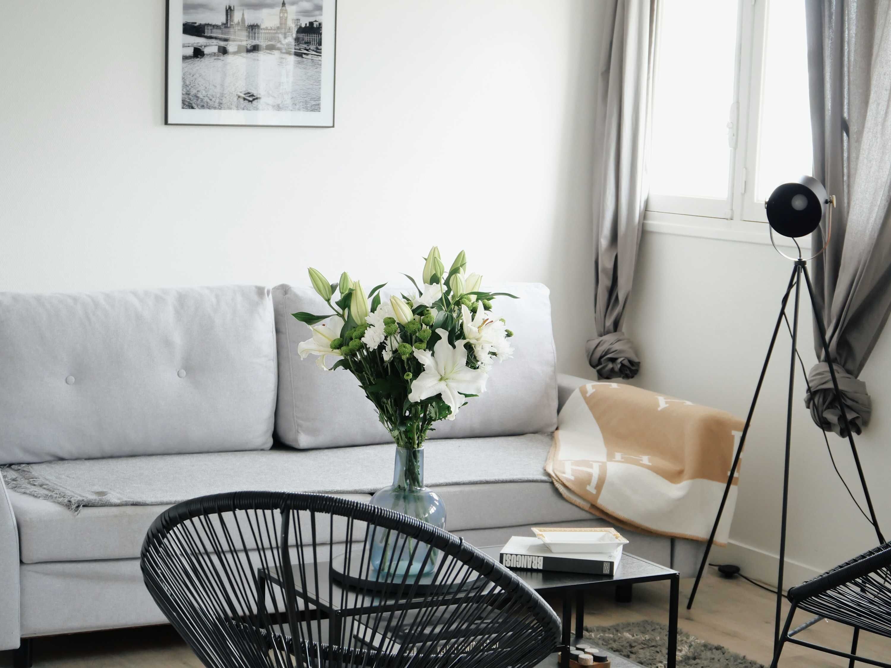 A modern living room with a grey sofa, a black wicker chair, a small table with a book, and a vase of flowers.