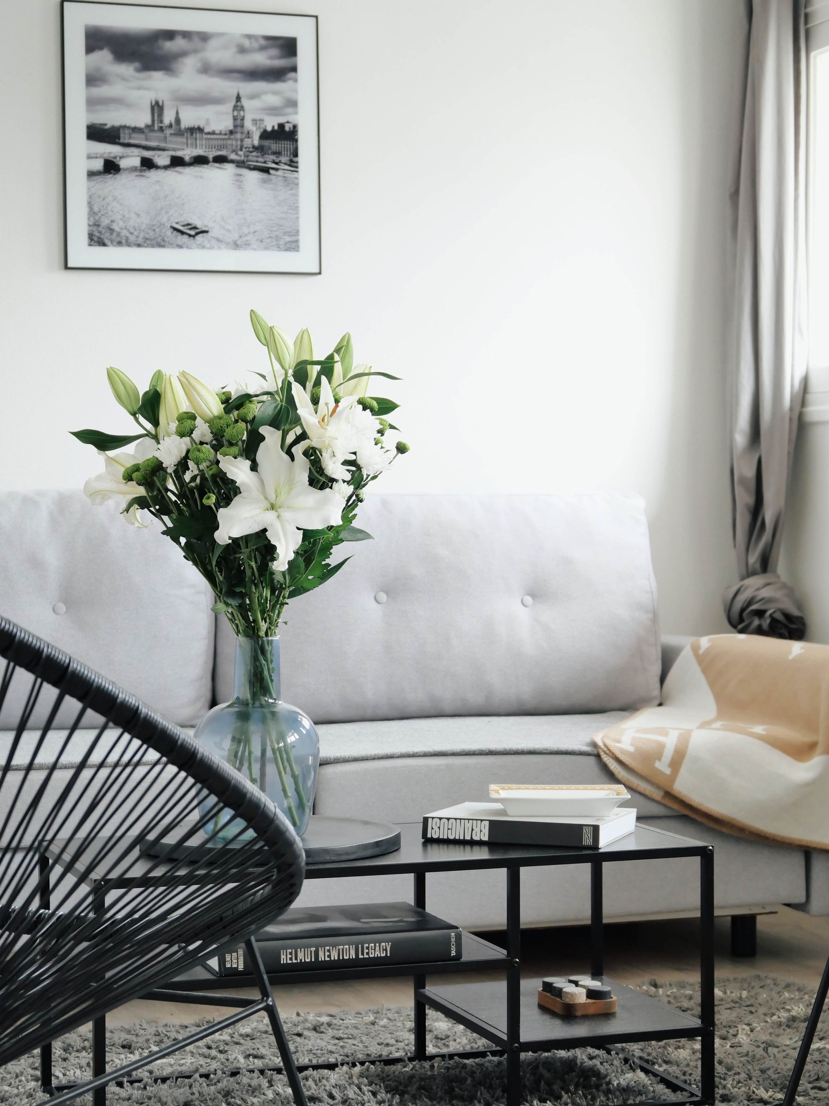 Un salon moderne avec un canapé gris, un fauteuil noir, un vase en verre contenant des fleurs blanches et un tableau encadré accroché au mur.