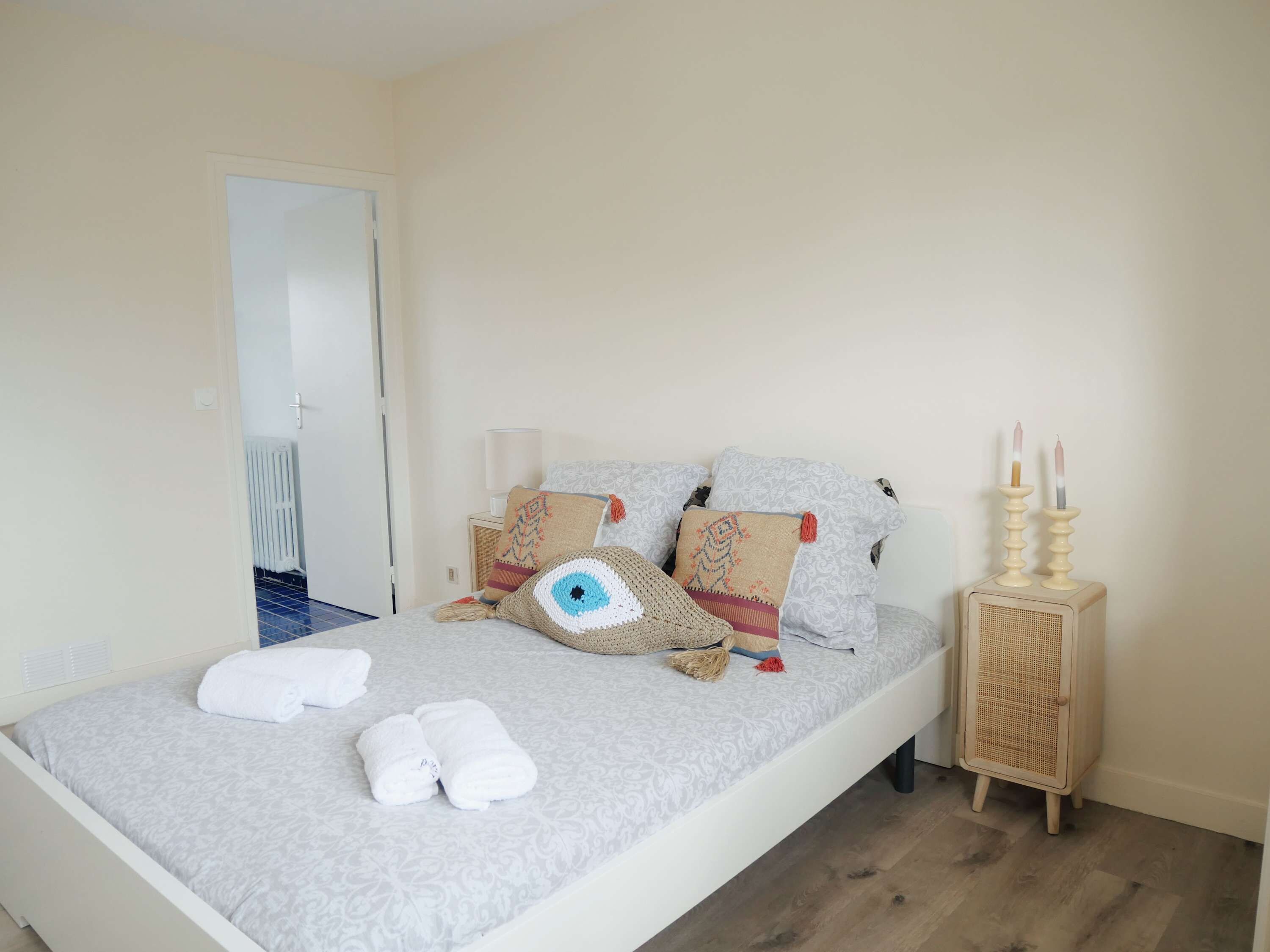 Ein ordentlich bezogenes Bett mit einer grauen, gemusterten Tagesdecke, zwei weißen Handtüchern und dekorativen Kissen mit Fransen. Ein Nachttisch aus Holz mit einem gewobenen Korb und zwei Kerzen steht neben dem Bett.