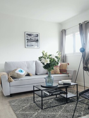 Ein modernes Wohnzimmer mit grauem Sofa, einem Couchtisch, einer Blumenvase, einem gerahmten Bild an der Wand und einem Fenster mit Vorhängen.
