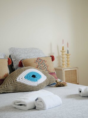 Ein ordentlich gemachtes Bett mit einem Kissen mit eingestricktem Augenmuster, einem aufgerollten weißen Handtuch und einem dekorativen Kerzenhalter mit Kerzen.
