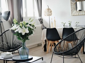 Ein Essbereich mit einem für zwei gedeckten Tisch, schwarzen Drahtstuhlen, einer Vase mit weißen Blumen und einer Stehlampe.