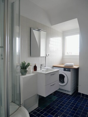 Ein modernes Badezimmer mit Duschkabine aus Glas, Waschbecken mit Spiegel darüber, integriertem Waschmaschinenanschluss und Fenster für natürliches Licht.