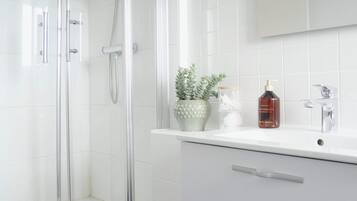 Um banheiro moderno com chuveiro em box de vidro, paredes com azulejos brancos, piso azul e pia branca com espelho, além de armário branco com gavetas.