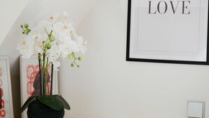 Eine weiße Wand mit einem gerahmten Bild des Wortes „LOVE“ und einer schwarzen Vase mit weißen Orchideen auf einer weißen Oberfläche.