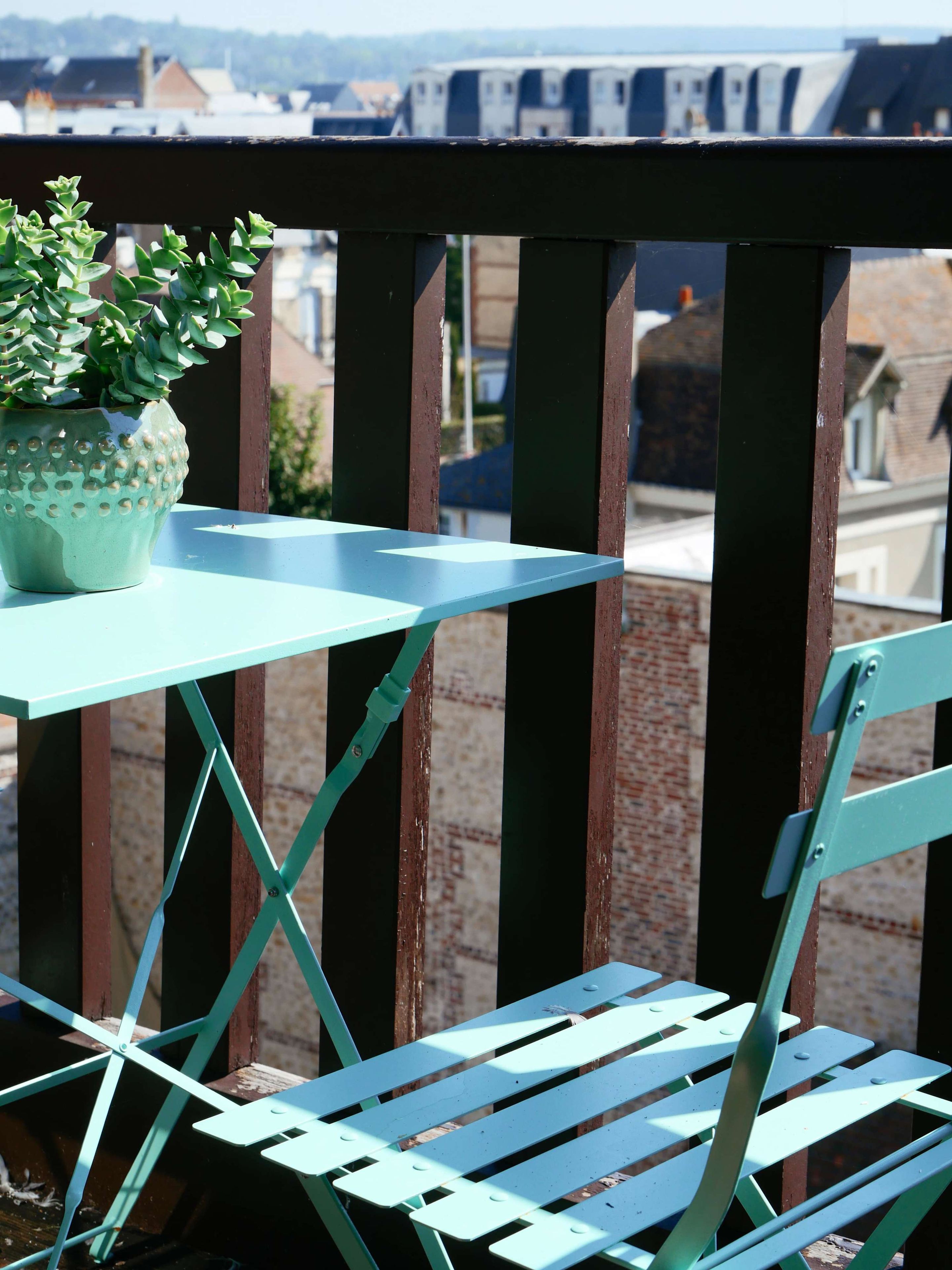 Un balcon avec une petite table et une chaise, un pot de fleurs et une vue sur les bâtiments en arrière-plan.