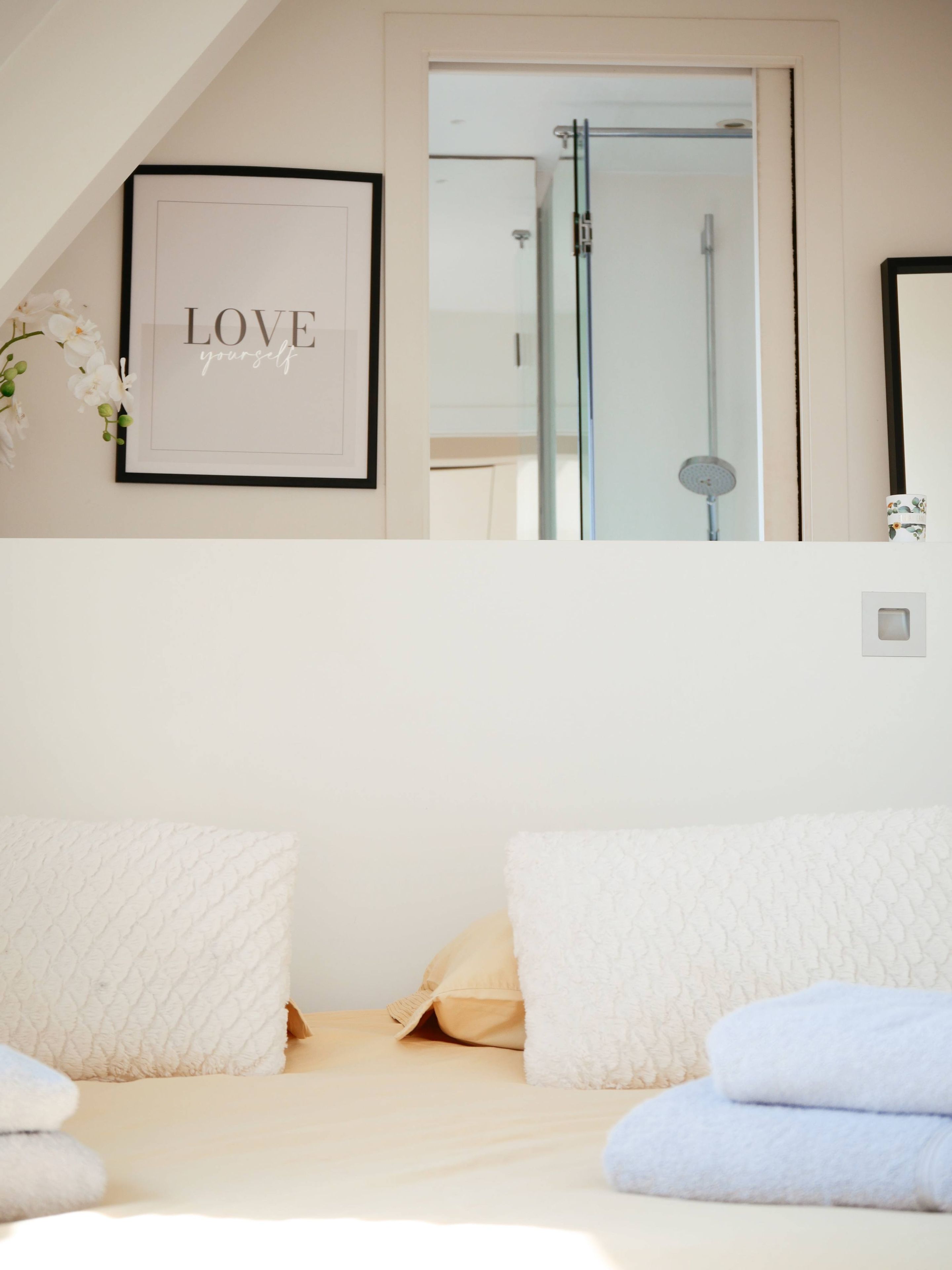Une chambre à coucher avec une citation inspirante encadrée au mur, un lit recouvert de draps blancs et une douche visible à travers une porte ouverte.