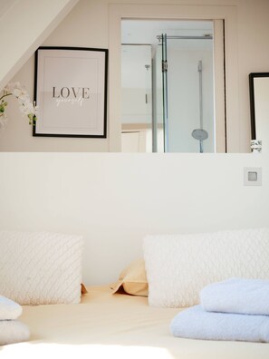 Un dormitorio con una cita inspiradora enmarcada, una cama con ropa de cama blanca y una ducha visible a través de una puerta abierta.