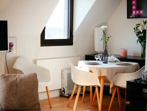 Et spiseområde med et rundt bord dækket til fire personer, hvide stole, en vase med blomster, en radiator og et vindue med sort ramme.