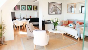 Ein modernes Wohnzimmer mit Sofa, Couchtisch und Sesseln, ein Essbereich mit Tisch und Stühlen sowie eine Küche im Hintergrund.