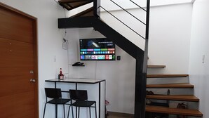 A compact room with a wooden door, a small table with two chairs, a TV mounted on the wall, and a staircase with wooden steps.