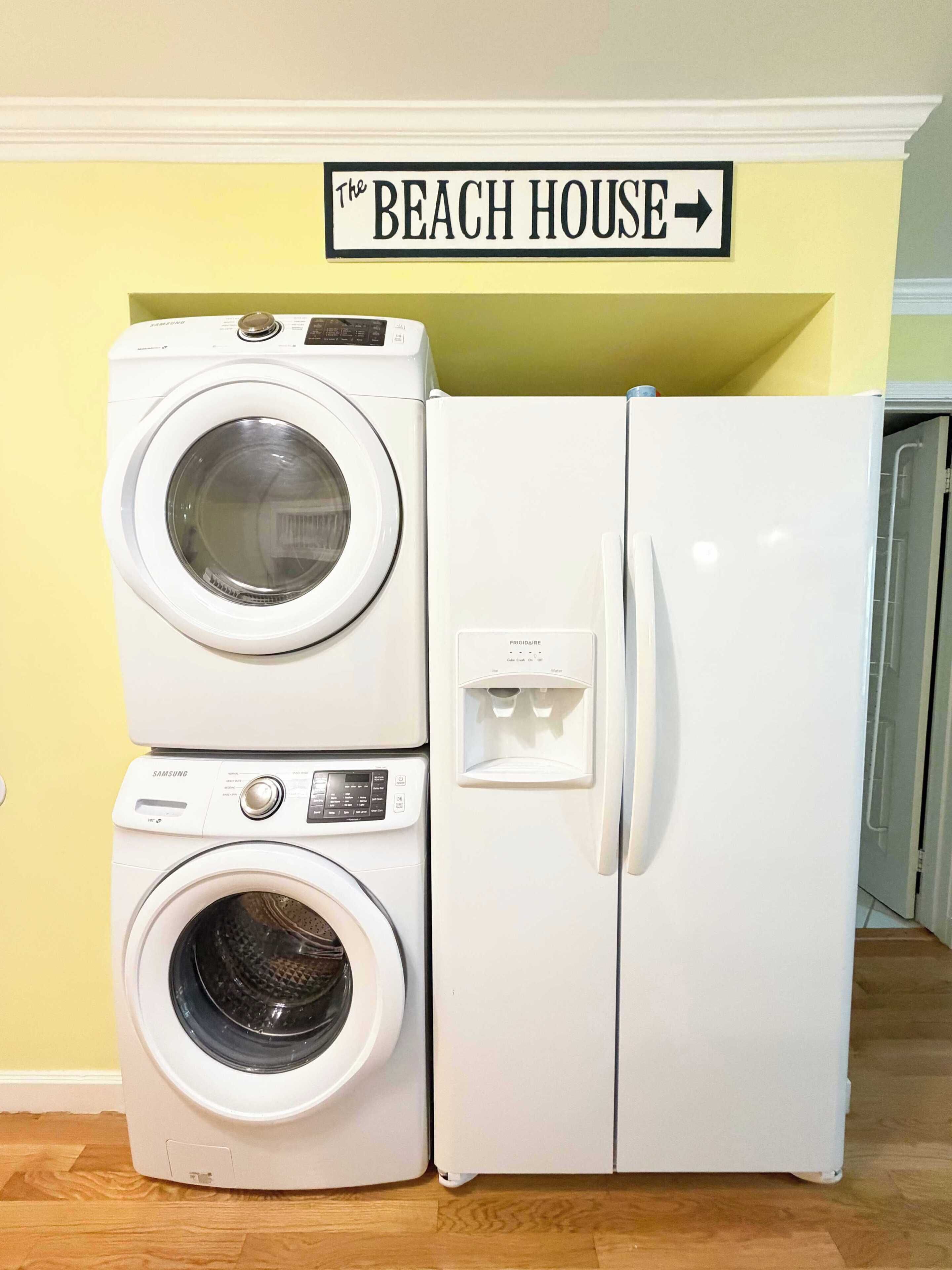 Un espace buanderie comprenant un lave-linge et un sèche-linge Samsung superposés, un réfrigérateur Frigidaire, et une pancarte au-dessus indiquant « THE BEACH HOUSE ».