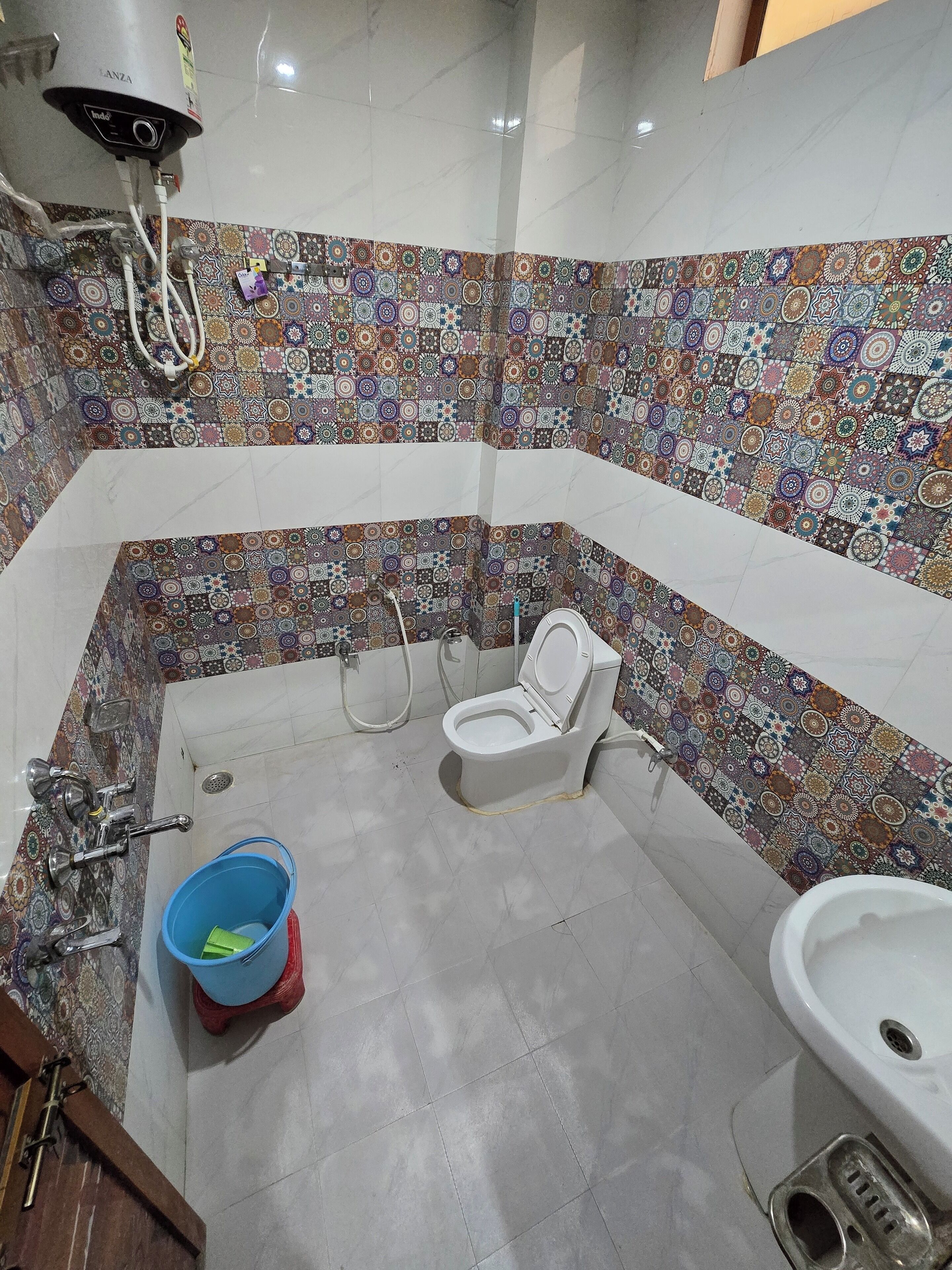 Et badeværelse med væg i flisemønster, toilet, bidet, håndvask og en blå spand.