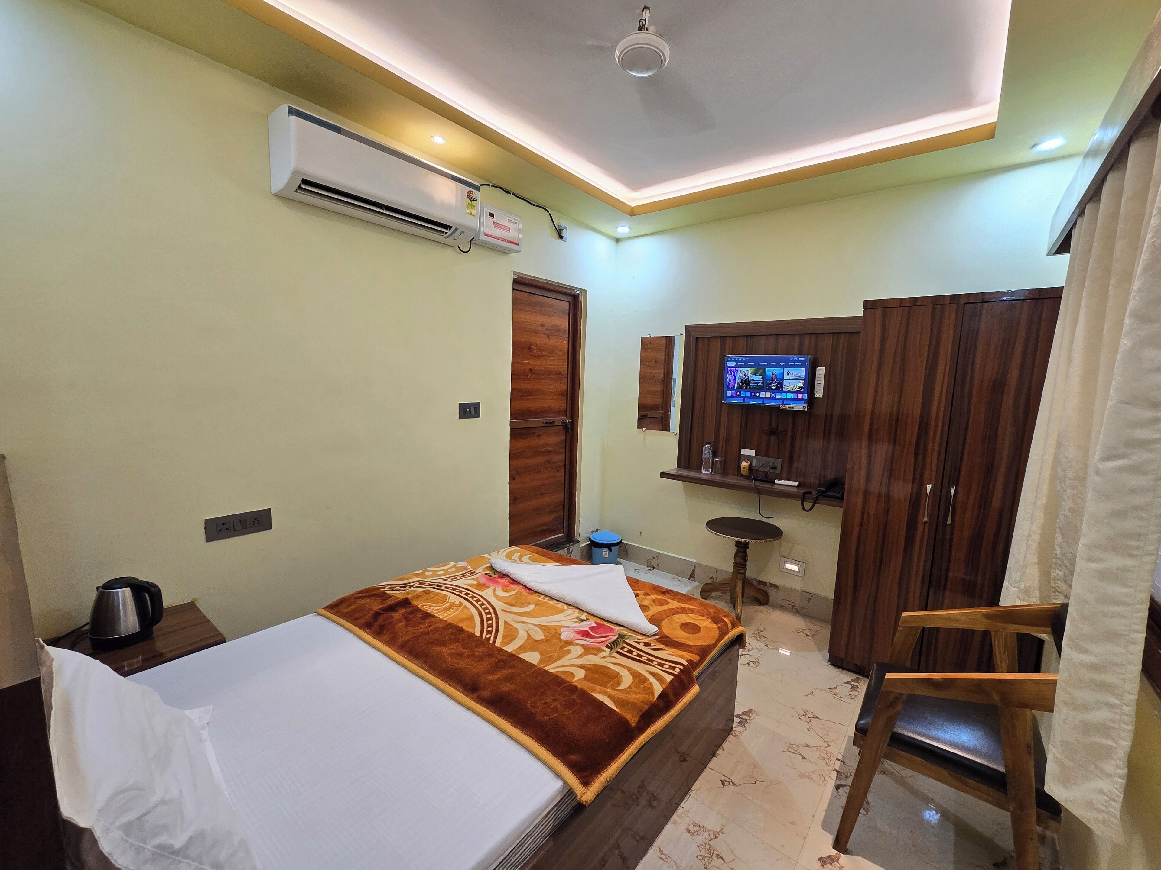 A hotel room with a bed, a TV mounted on the wall, a wooden wardrobe, a small table, and a chair.
