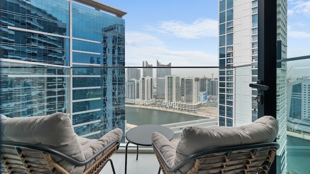A balcony with wicker chairs and a small table, overlooking a cityscape with modern buildings and a body of water. - Waves - Chic High-Floor Studio Retreat with Canal Views (Dubai)