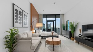 A modern hotel room with a sofa, a round coffee table, a TV, and a large window offering a city view. - Waves - Chic High-Floor Studio Retreat with Canal Views (Dubai)