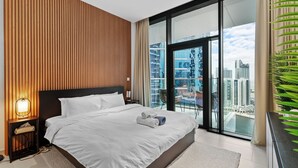 A modern hotel room with a large bed, a nightstand, a lamp, and a view of the city through floor-to-ceiling windows. - Waves - Chic High-Floor Studio Retreat with Canal Views (Dubai)