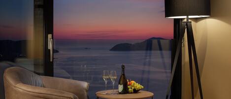 A room with a view of the sea at sunset, a table with a bottle and glasses, and a lamp.