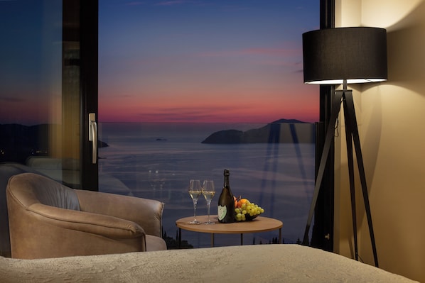 Une pièce avec vue sur la mer au coucher du soleil, une table avec une bouteille et des verres, et une lampe.