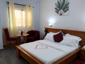 Hotellihuone, jossa on sänky, tuoli, pieni pöytä, lamppu ja taulu seinällä.