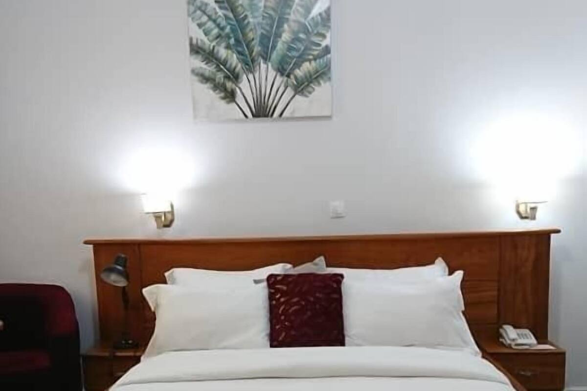 Ein ordentlich bezogenes Bett mit weißen Leinentüchern und einem roten Zierkissen, ein hölzernes Kopfteil sowie ein an der Wand befestigtes Kunstwerk darüber.