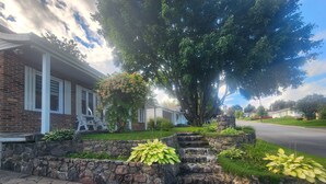 Ein Wohnhaus mit einer Steinstützmauer, einem kleinen Garten mit Pflanzen und einer gepflasterten Auffahrt, die zu einem Haus mit Veranda führt.