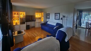 Ein Wohnzimmer mit blauem Sofa, einem Couchtisch und einem Fernseher. Es gibt eine Küchenzeile mit Schränken und einem Esstisch.