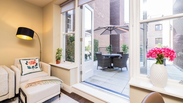 A cozy room with a sofa, a small table, and a window overlooking a patio with outdoor furniture and plants.