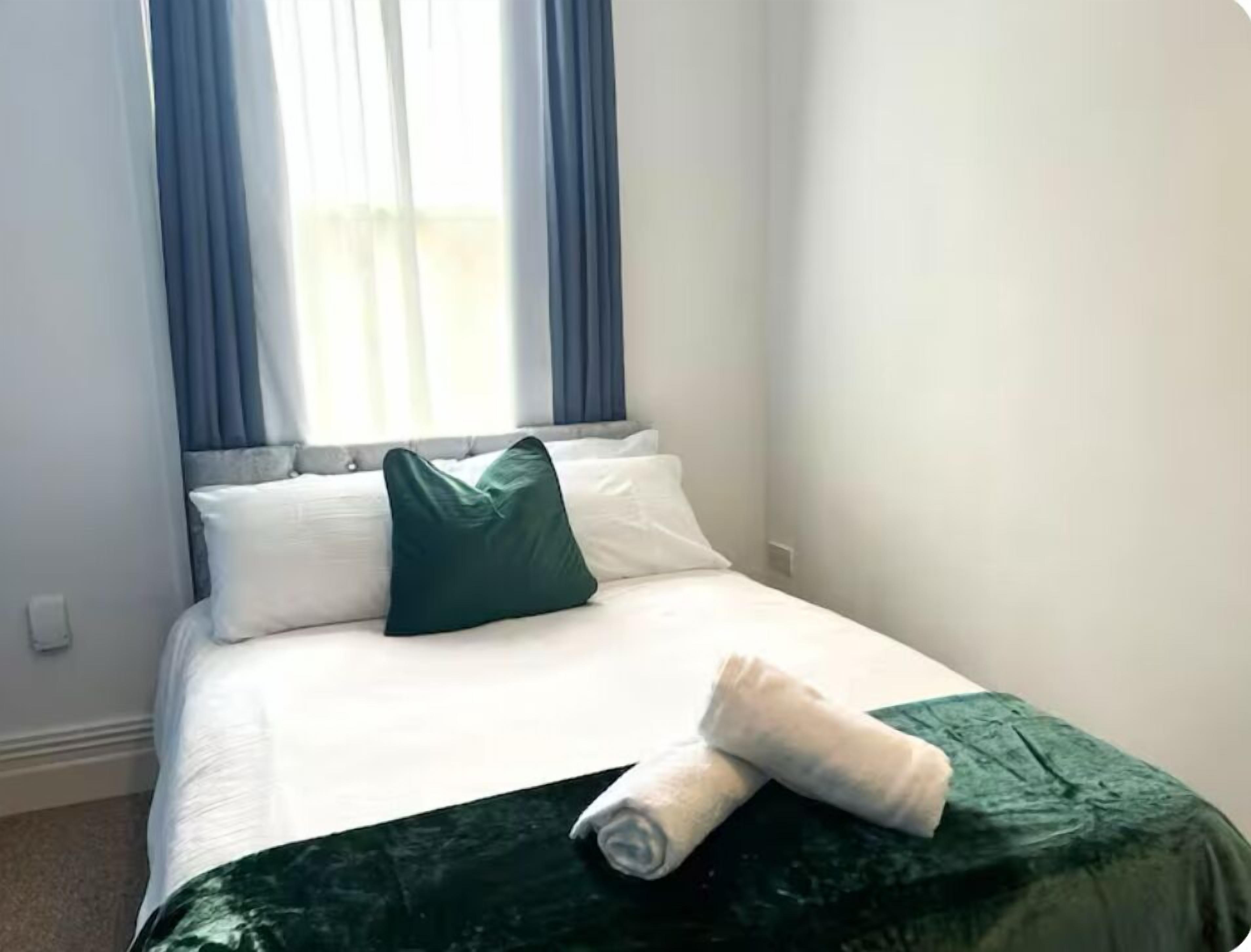 Una cama bien hecha con sábanas blancas y un cojín verde, una toalla blanca enrollada sobre la cama, y una manta verde oscuro.
