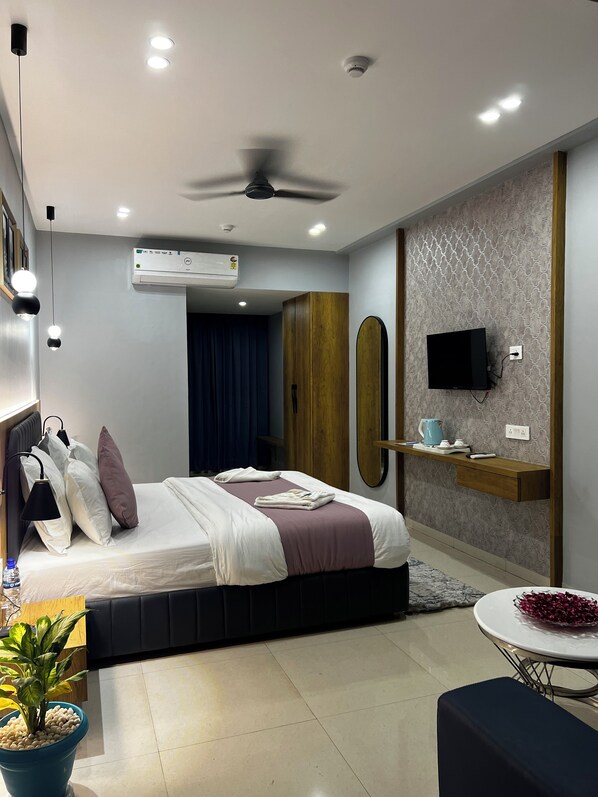 A modern bedroom with a bed, bedside lamps, a ceiling fan, an air conditioning unit, a TV, a small table with a kettle, and a potted plant.