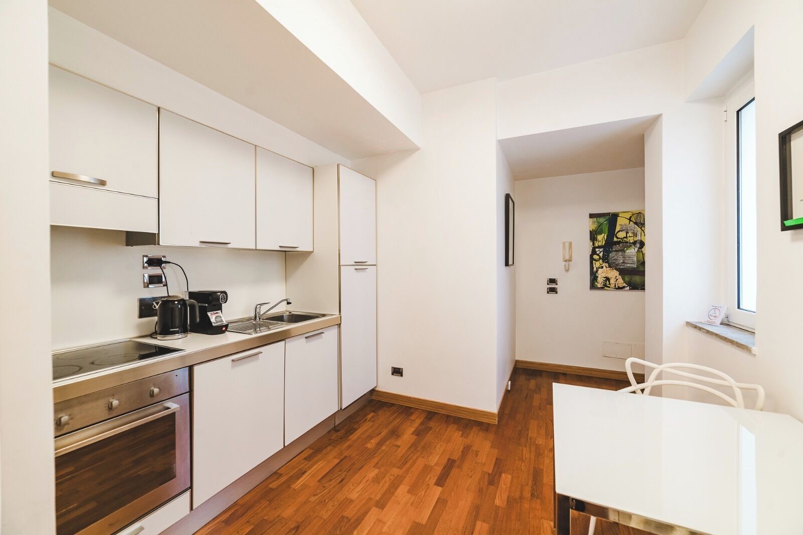 Una cocina compacta con armarios blancos, cafetera y lavamanos. Cuenta con un comedor con mesa y sillas blancas.