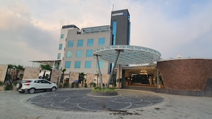 A modern hotel with a central circular entrance, a tall building with a 'Sapphire Blue' sign, and a parking area with a white car. - Sapphire Blue (Bhopal)