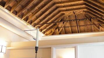 A room with a high, wooden ceiling, a dining table with a bowl of lemons, a lamp, and a bed with a TV.