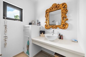 A bathroom with a white sink, a gold-framed mirror, and a window with a view of palm trees. - Charming apartment with AC in living, garden and walking distance to the ocean! (Kralendijk)