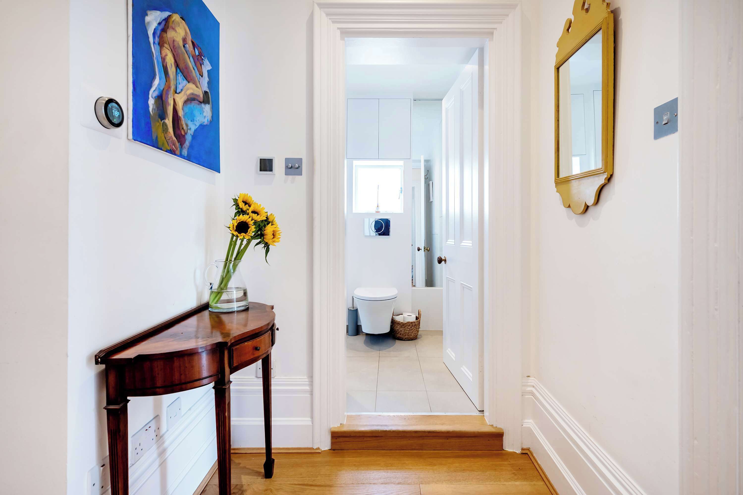 A hallway with a wooden console table, a vase with sunflowers, a painting, a digital thermostat, and a mirror.