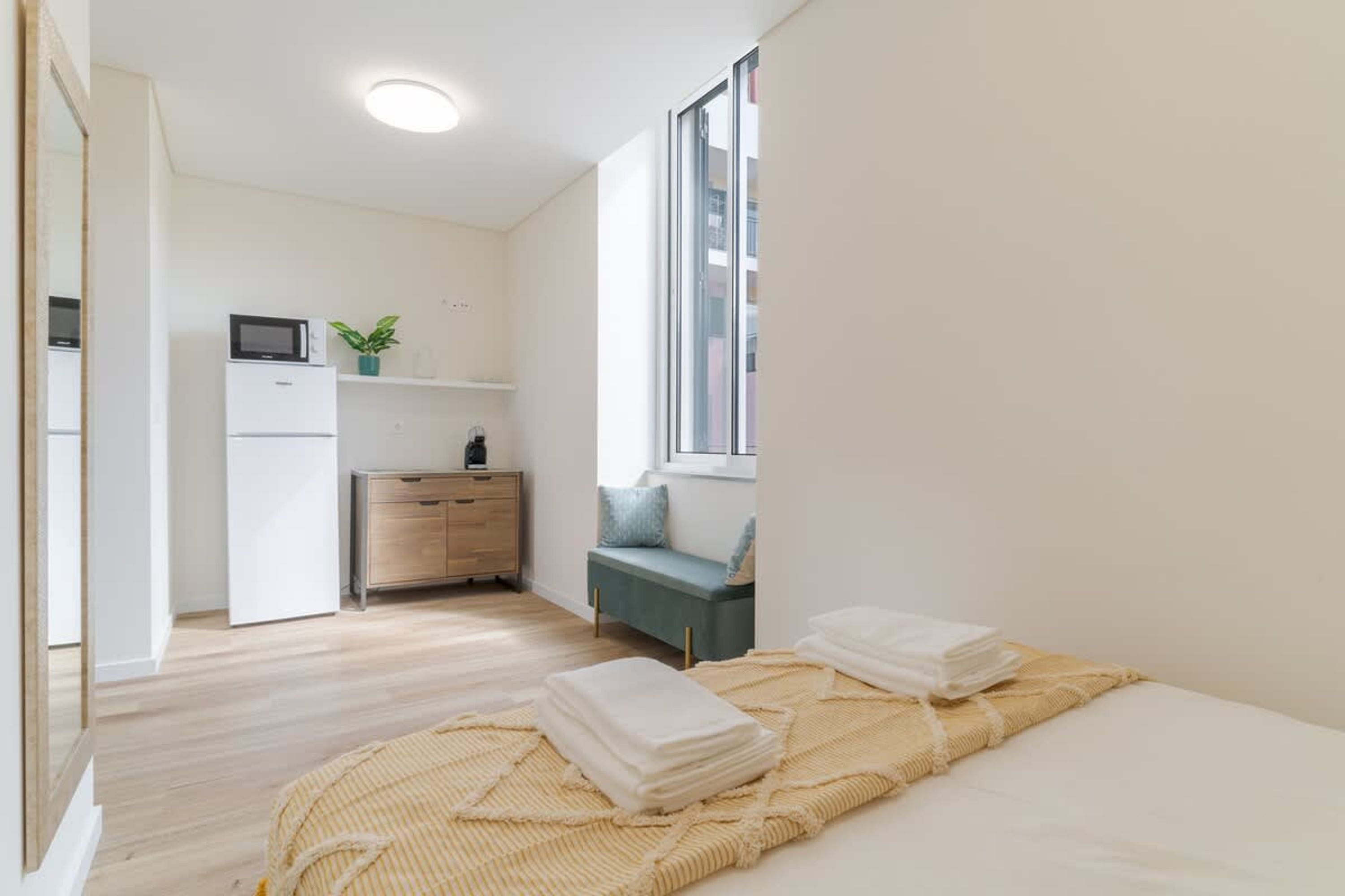 A modern, minimalist bedroom with a bed, a bedside table, a small desk, a microwave, and a window.