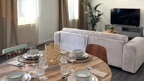 Un comedor moderno con una mesa redonda dispuesta para cuatro personas, un sofá al fondo y un televisor sobre un mueble bajo.