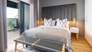 A modern bedroom with a large bed, two bedside lamps, a bench, and a view of the city through a balcony. - Gorgeous One Bedroom Oceans (uMhlanga Rocks, KwaZulu-Natal)