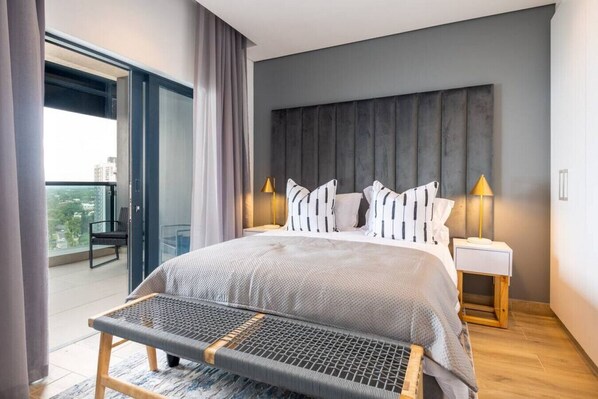 A modern bedroom with a large bed, two bedside lamps, a bench, and a view of the city through a balcony.