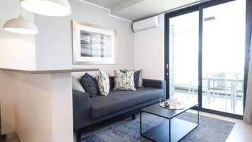 A modern living room with a dark grey sofa, patterned cushions, a glass coffee table, and a large window offering a view of the outdoors.