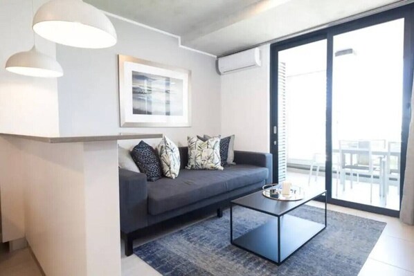 A modern living room with a dark grey sofa, patterned cushions, a glass coffee table, and a large window offering a view of the outdoors.